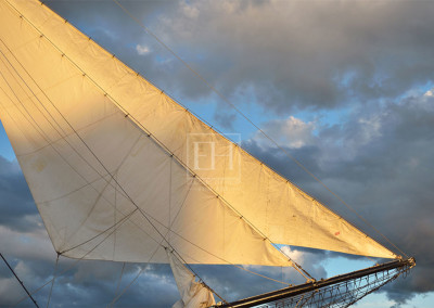Close up photographic image of billowing white sails of a tall ship glowing golden in the light of the setting sun.
