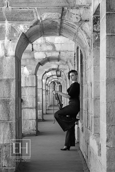 A violinist dressed in black with violin in hand standing in upper balcony corridor of Fort Henry in Kingston, Ontario as photographed by Elizabeth Hesp